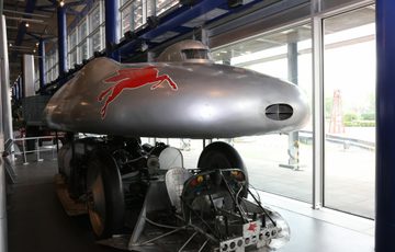 Silver coloured motorvehicle with a red winged horse portrayed on its side. The motor vehicle is long and thin.