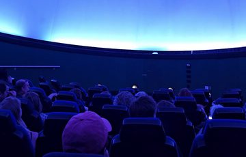 A planetarium full of people looking the blue screen above.