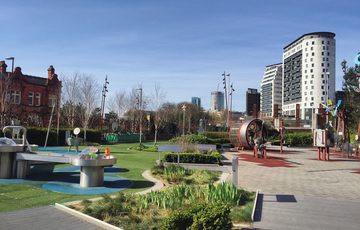 A science garden with trees, plants and science exhibits and two buildings in the distance.