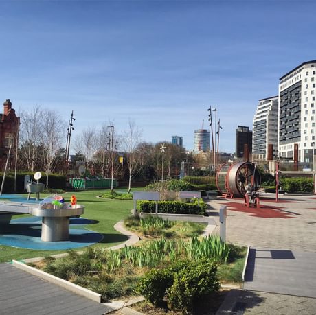 A science garden with trees, plants and science exhibits and two buildings in the distance.