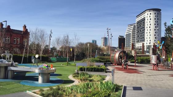 A science garden with trees, plants and science exhibits and two buildings in the distance.