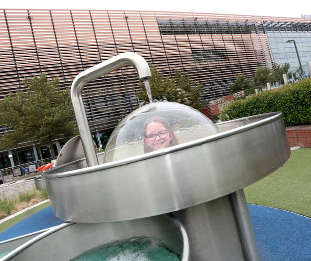 A child using a science exhibition in a garden. She is a under a plastic dome and water pouring over the top of the dome.