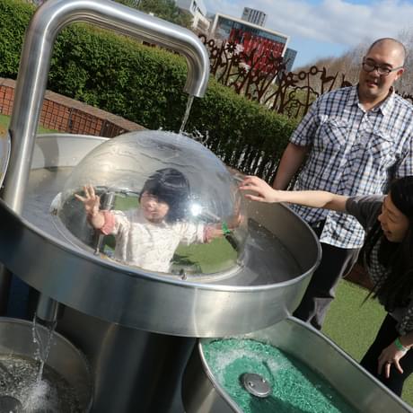 Two adults watching a child in a plastic dome with water running over the top of it.
