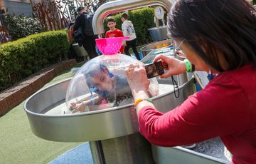 A child in a plastic dome with water running over the top. An adult is taking a photo.