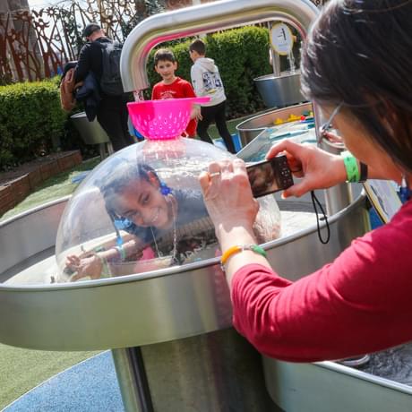 A child in a plastic dome with water running over the top. An adult is taking a photo.