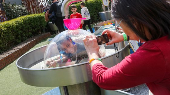 A child in a plastic dome with water running over the top. An adult is taking a photo.