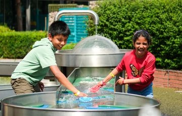 Two children play joyfully with water and colourful toys at an outdoor fountain.