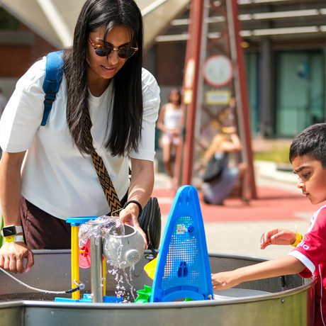 Woman and boy engaging with water play exhibit outdoors on a sunny day.
