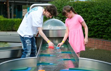 A person and child play with floating toys in an outdoor water play area.