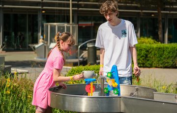 An adult and child play with a water activity exhibit outdoors on a sunny day.