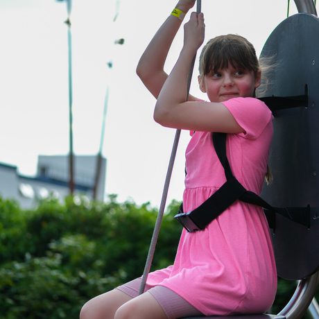 Girl in pink dress enjoying hoisted chair ride outdoors with greenery and buildings in the background.