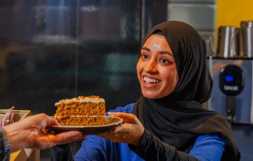 A staff member who is smiling hands a slice of cake to a visitor.