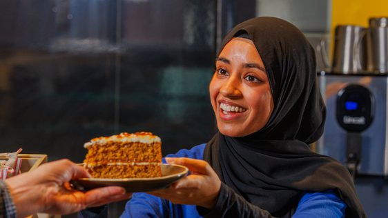 A staff member who is smiling hands a slice of cake to a visitor.