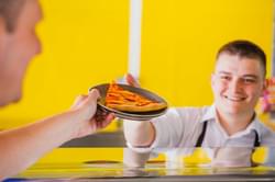 A smiling member of staff serving a slice of pizza to a customer over the counter.