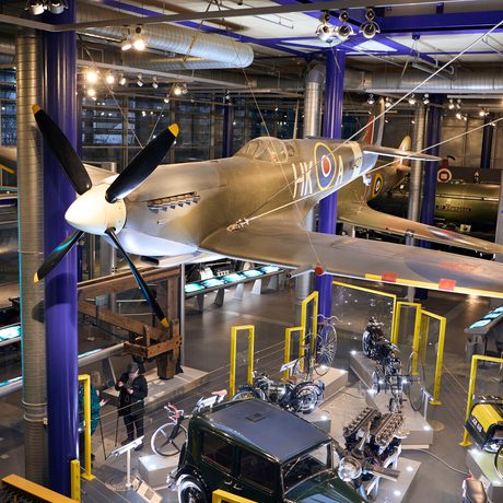 A WWII military aircraft suspended above classic vehicles and engines in a museum exhibit.