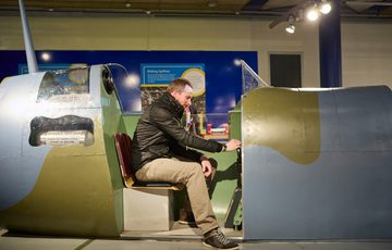 Man sitting inside a replica spitfire cockpit, examining the controls.