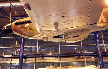 Three people observe a The Railton Land Speed car with and the Hawker Hurricane aeroplane suspended above in a museum gallery.