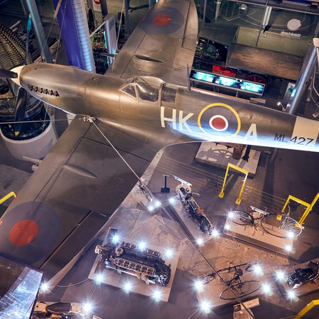 World War II Spitfire plane displayed suspended above illuminated exhibits, engines and historic vehicles.