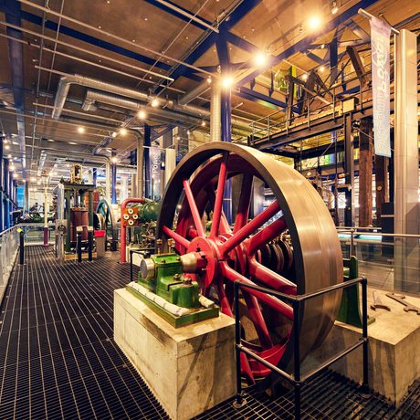 Large red industrial flywheel with green machinery and metal grating flooring inside museum setting.