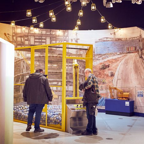 Two people playing with an interactive digger exhibit within a yellow cage filled with colourful balls.