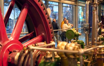 Large red industrial flywheel with complex machinery, people observing behind glass barrier.