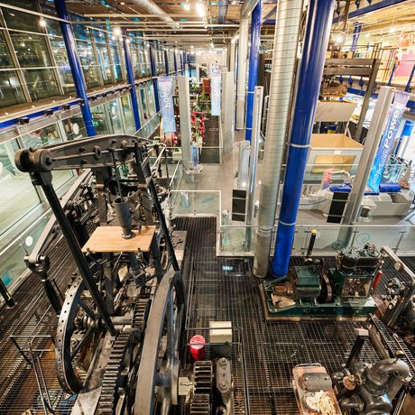 Large, industrial machinery displayed inside a modern, glass-walled museum with visitors and exhibits.