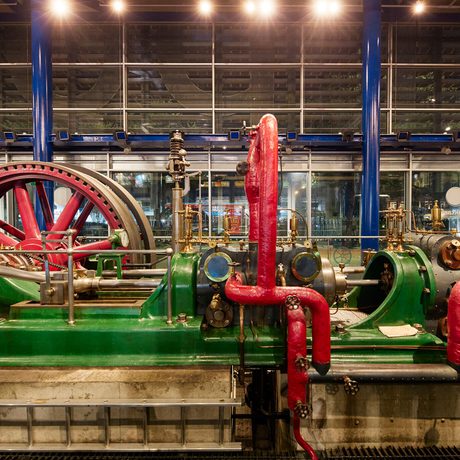Large industrial steam engine with red pipes and green machinery in a well-lit factory setting.