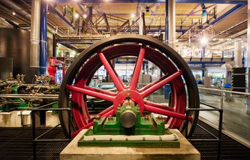 Large red industrial flywheel inside a museum with metal pipes and machinery surrounding it.