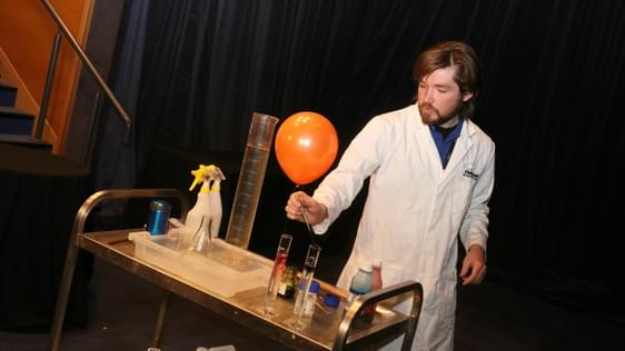 A man dressed in a white coat is giving a fun science demonstration. In front of him is an orange balloon and two very large test tubes holding liquid.