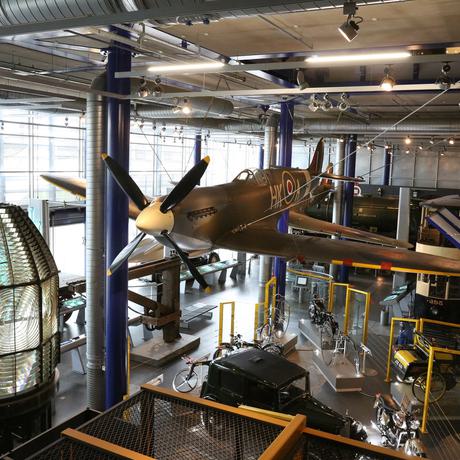 Large exhibition space containing a Spitfire plane suspended over vintage cars and bikes