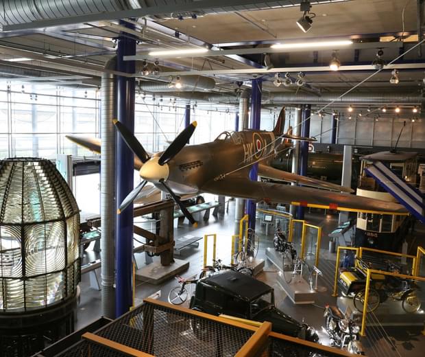Large exhibition space containing a Spitfire plane suspended over vintage cars and bikes