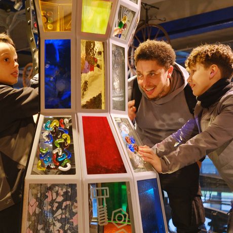 An adult and two children interact excitedly with a colourful, illuminated, glazed interactive exhibit.