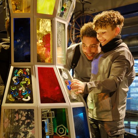 An adult and two children interact excitedly with a colourful, illuminated, glazed interactive exhibit.