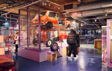 A vintage mini car displayed elevated in an interactive museum with visitors exploring exhibits.