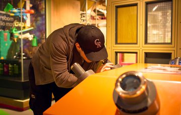Person wearing a black cap peers into a microscope device at an interactive museum display.