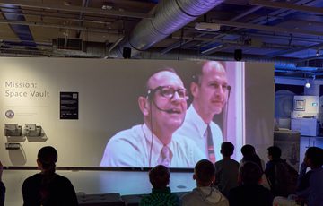 Back of heads of a group of people sitting watching a large video on display.