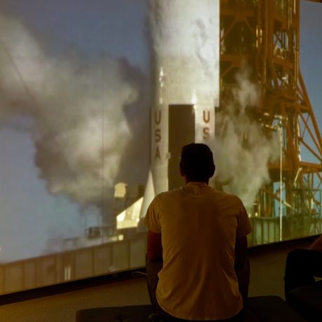 Two people sitting in front of large screen watching a rocket launch.