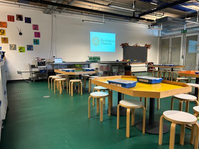 A studio classroom with stools, tables with lips and green floor with a LEGO clock on the wall.