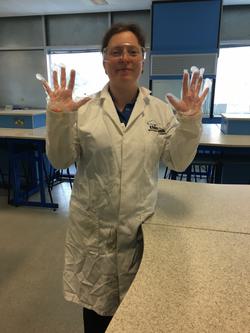 Member of staff with their hands up while wearing gloves, a lab coat and protective goggles.