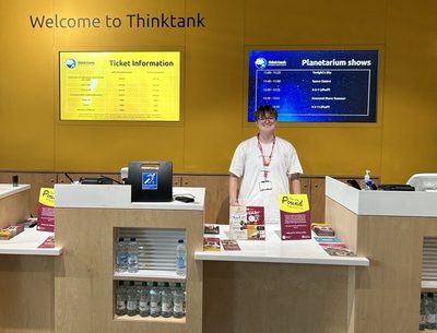 A staff member wearing a pink lanyard is standing behind the ticket desk.