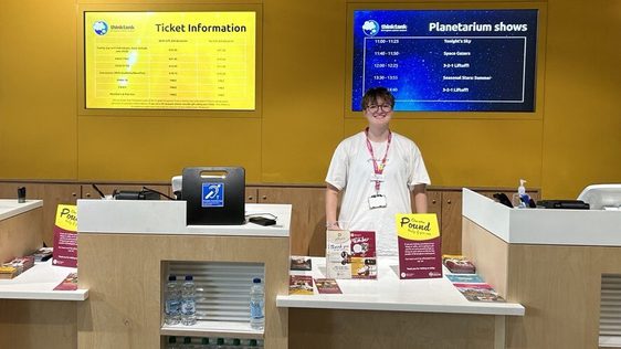 A staff member wearing a pink lanyard is standing behind the ticket desk.