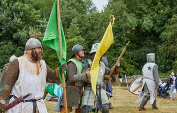 Three re-enactors dressed as Medieval knights holding yellow and green flags with trees in the background.