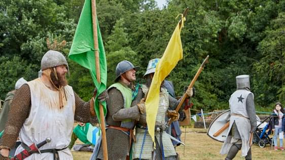 Three re-enactors dressed as Medieval knights holding yellow and green flags with trees in the background.