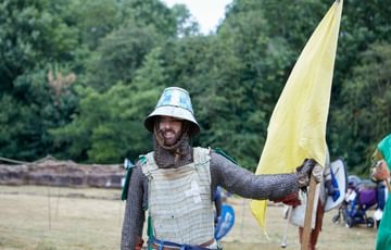A re-enactor dressed as a medieval knight holding a large yellow flag, trees and grass are in the background. s