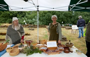 Two women dressed in medieval clothes with a selection of medieval foods and baskets on a table in front of them.