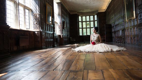 A bride sitting on an old wooden floor in a the long gallery at Aston Hall. The wood panelling and windows surround her. Her dress is laid out and she holds a red bouquet of flowers while looking to the side.