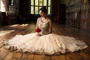 Close up of a bride sitting on the wooden floor in the Long Gallery. She is looking down and holding a red bunch of flowers, with her dress laid out around her.