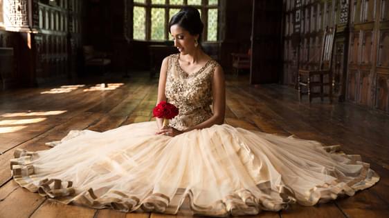 Close up of a bride sitting on the wooden floor in the Long Gallery. She is looking down and holding a red bunch of flowers, with her dress laid out around her.