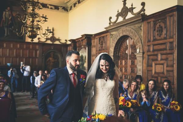 A bride and groom getting married in the Great Hall.  The bride is looking down smiling. The room has wood paneled walls and their guests are seated behind them.