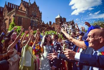 A bride and groom outside sharing a kiss, with a crowd scattering confetti over them. Aston Hall is in the background